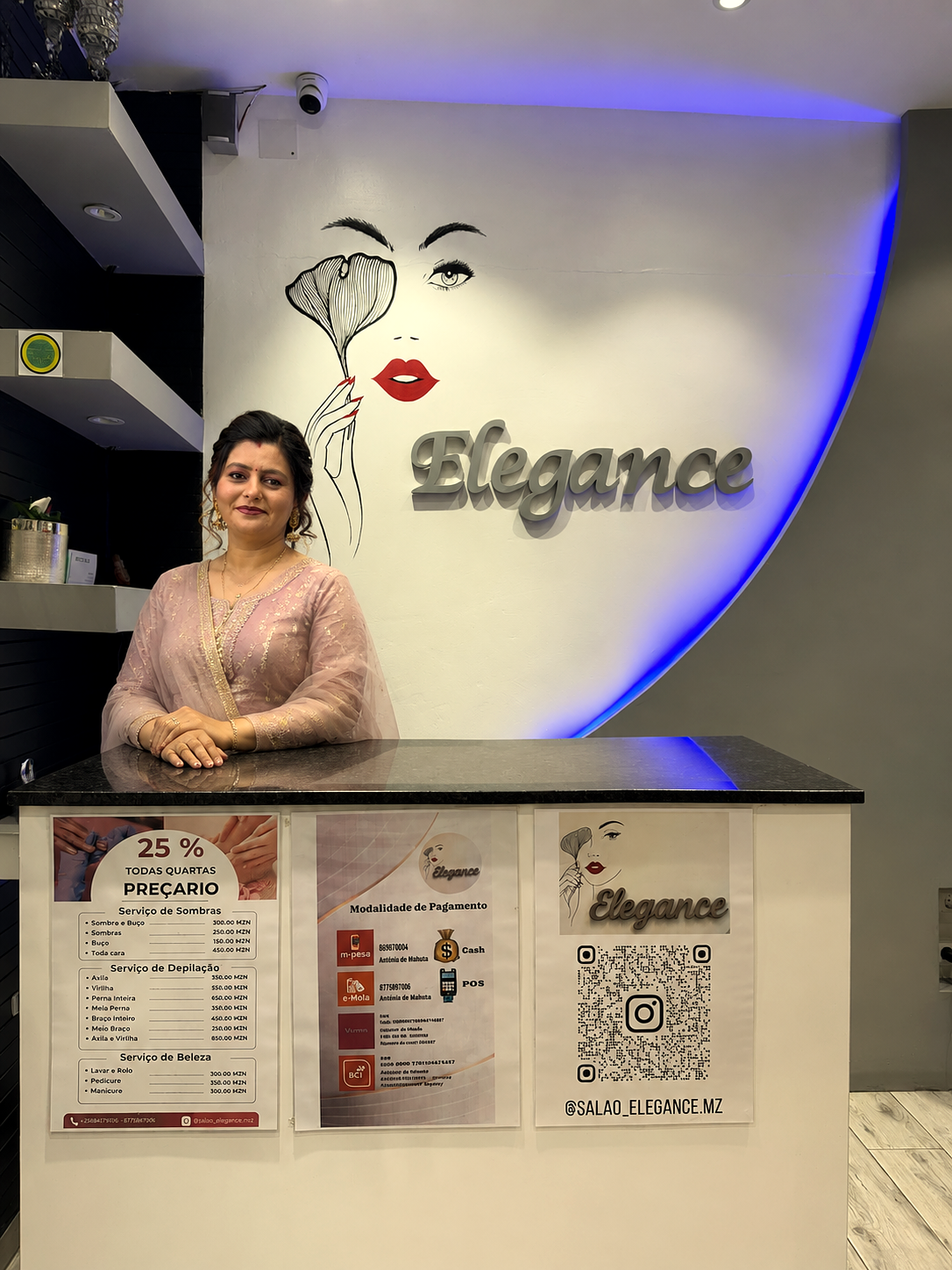 Founder standing behind the Salão Elegance reception desk with the Elegance wall logo and salon artwork visible